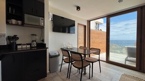 Eine moderne Küche mit dunklen Schränken, einer eingebauten Mikrowelle und einem Mixer auf der Arbeitsfläche. Ein Esstisch mit vier Stühlen und ein kleiner Mülleimer sind ebenfalls vorhanden. Ein großes Fenster bietet Meerblick.