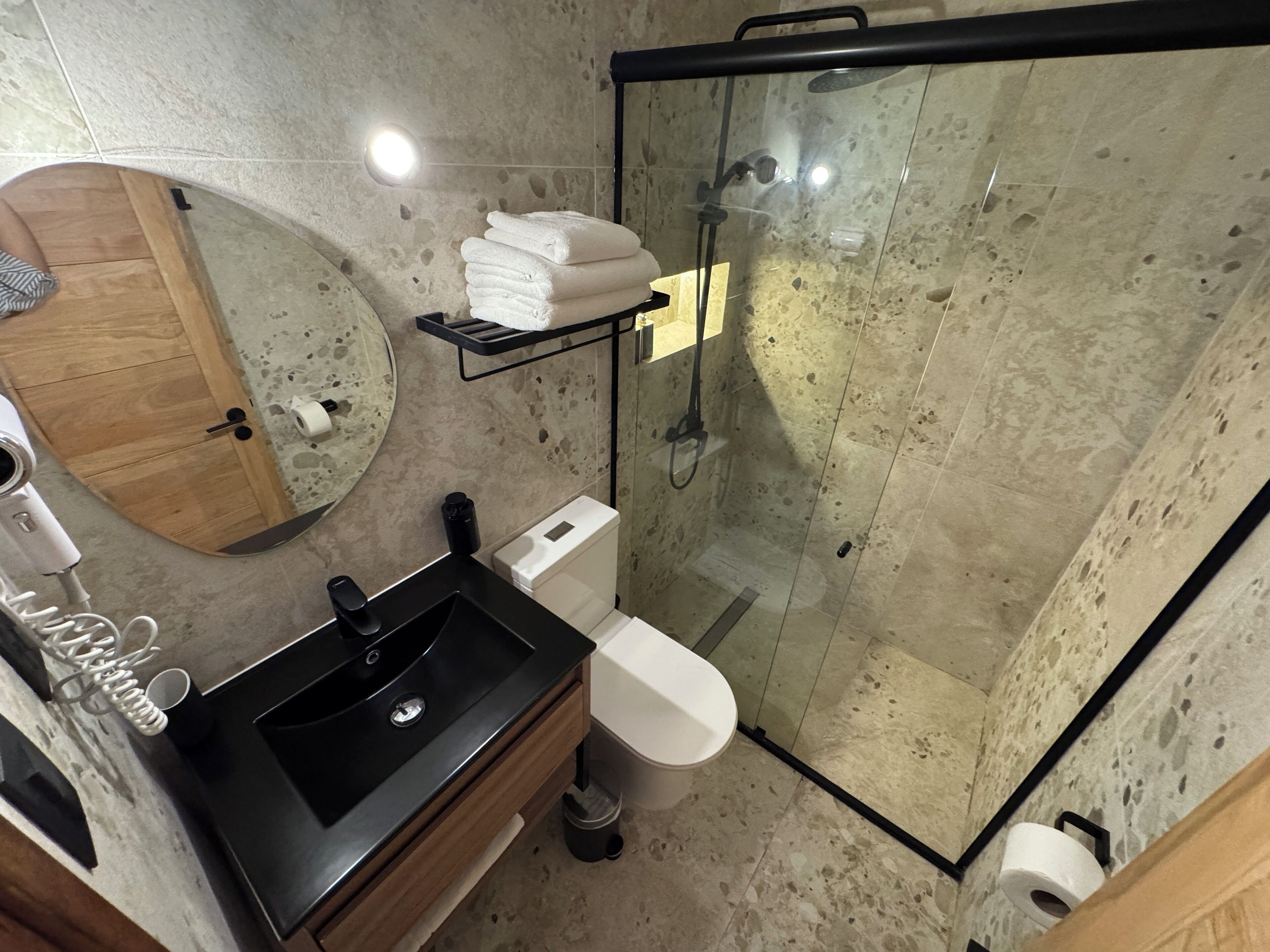 Un baño moderno con ducha envuelta en vidrio, lavamanos negro, tocador de madera y un perchero para toallas.
