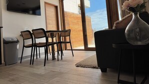 Ein Wohnzimmer mit an der Wand montiertem Fernseher, einem Essbereich mit Stühlen und Tisch, einer Couch, einer Vase mit Blumen und einer Glasschiebetür, die zu einem Außenbereich führt.