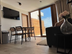 A living room with a TV mounted on the wall, a dining area with chairs and a table, a sofa, a vase with flowers, and a sliding glass door leading to an outdoor area.