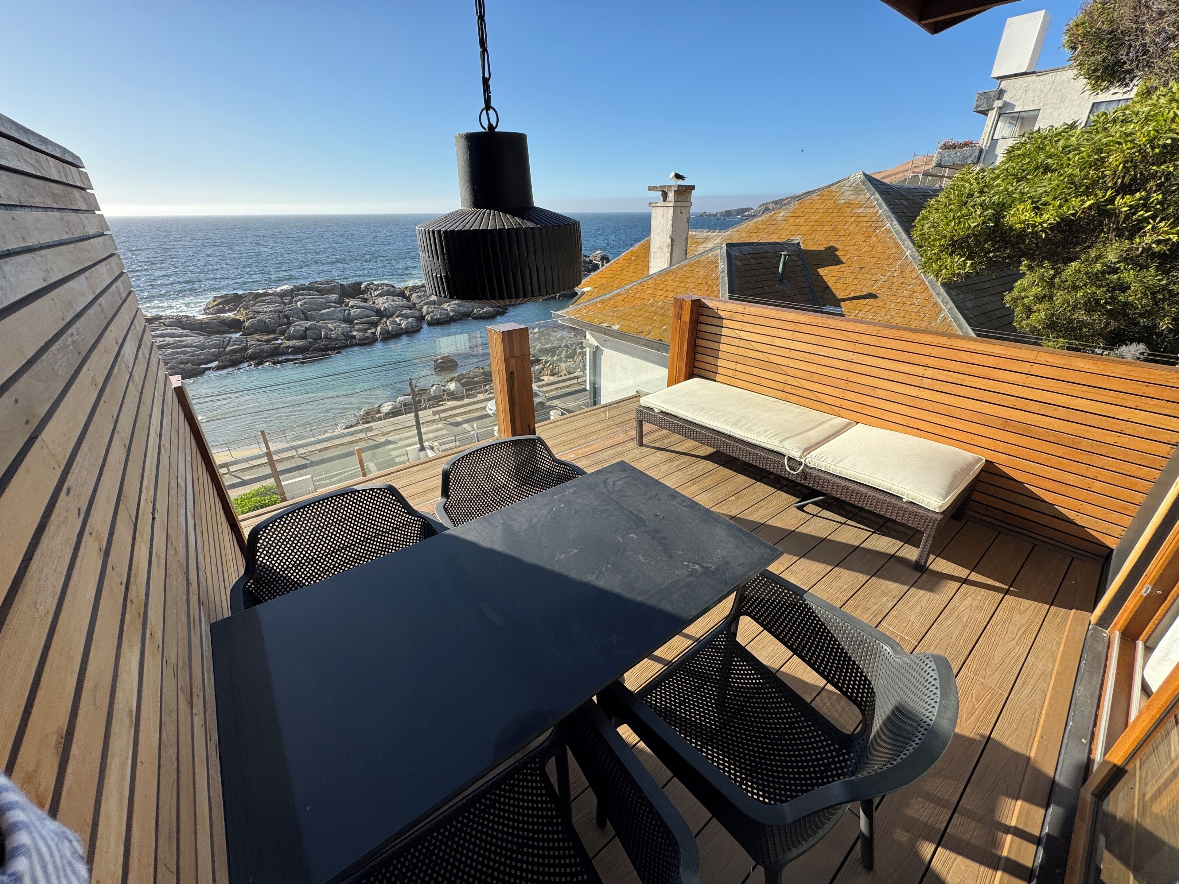 A wooden deck with a table and chairs, overlooking a rocky coastline and the sea.