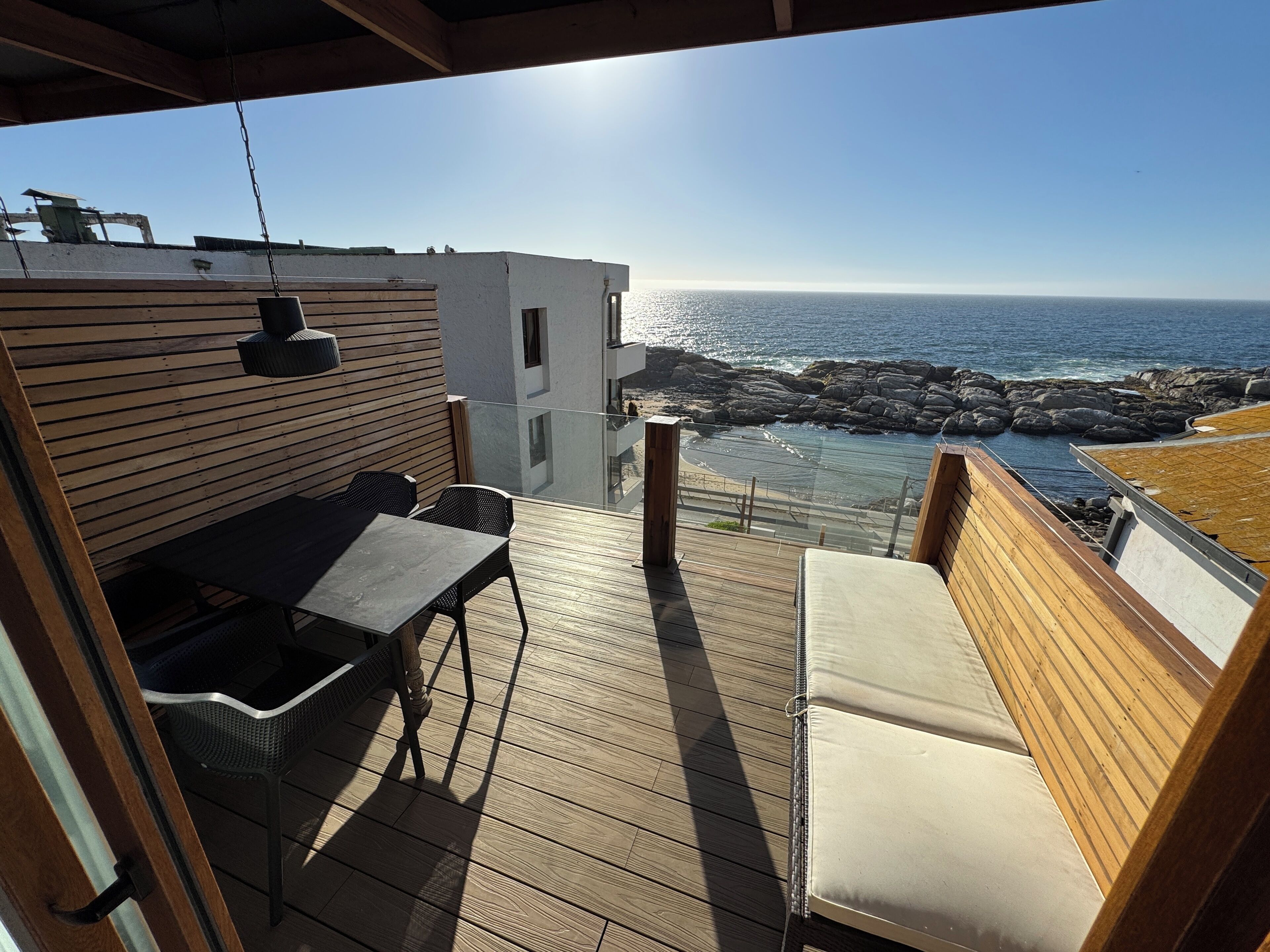 Een houten terras met tafel en stoelen, met uitzicht op een rotsachtige kustlijn en de zee.