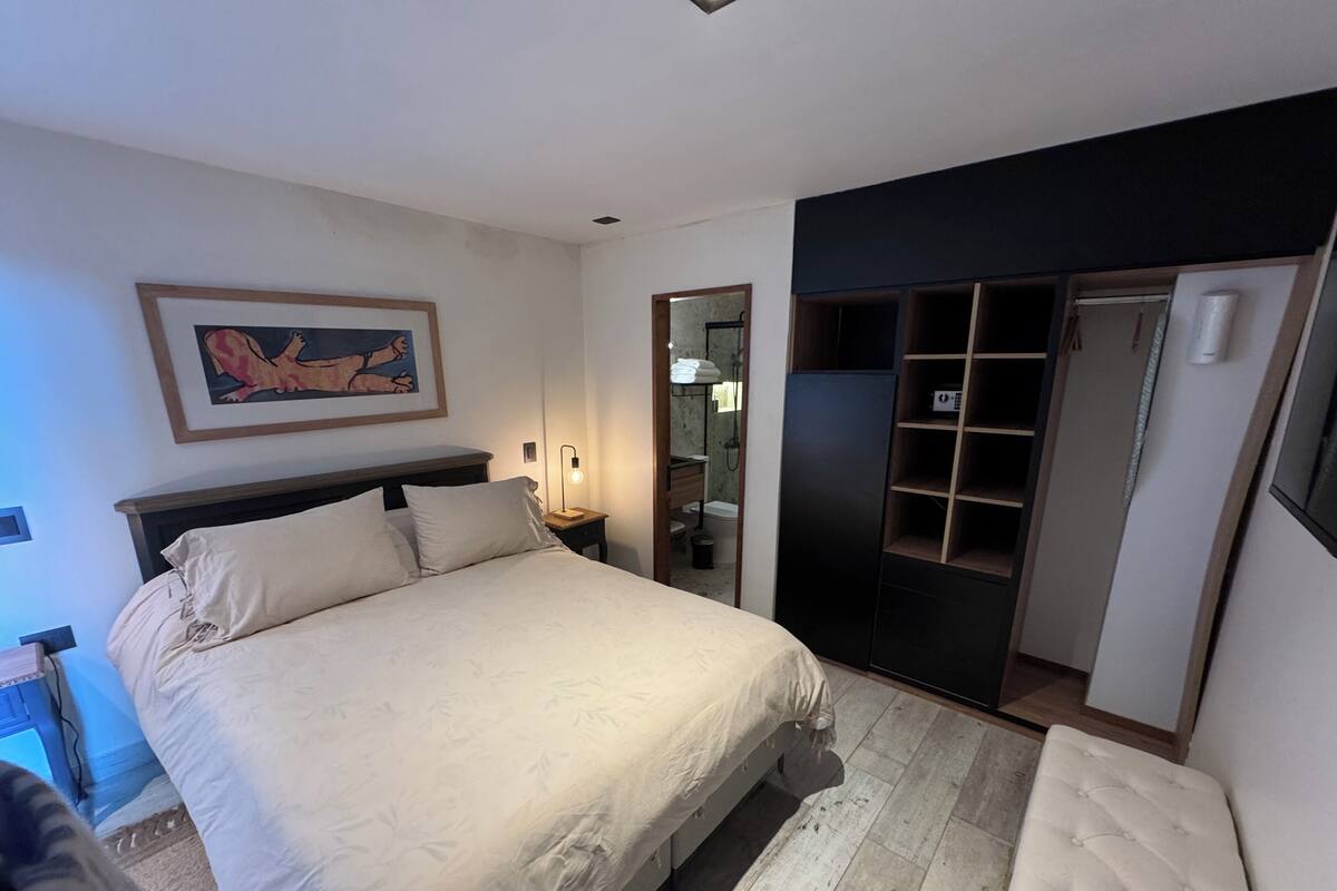 Un dormitorio con cama, mesita de noche, lámpara, armario y un cuadro en la pared.