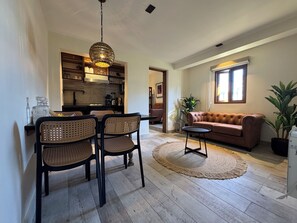 A living room with a brown sofa, two chairs, a coffee table, a dining area with a table and chairs, a kitchen in the background, and a large window with natural light.