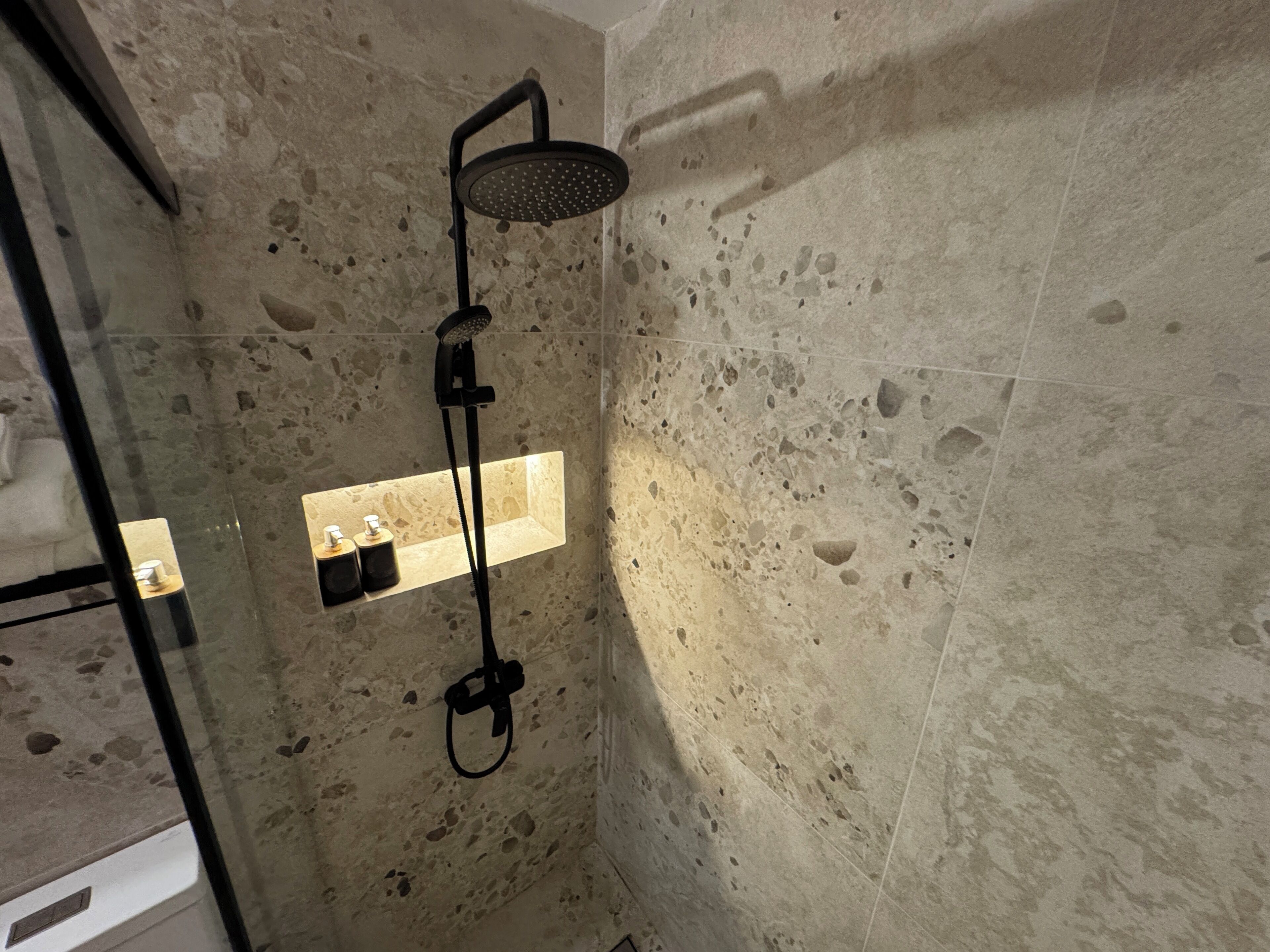 A shower with a handheld showerhead and a wall-mounted rain showerhead. There is a shelf with two bottles and a soap dispenser.