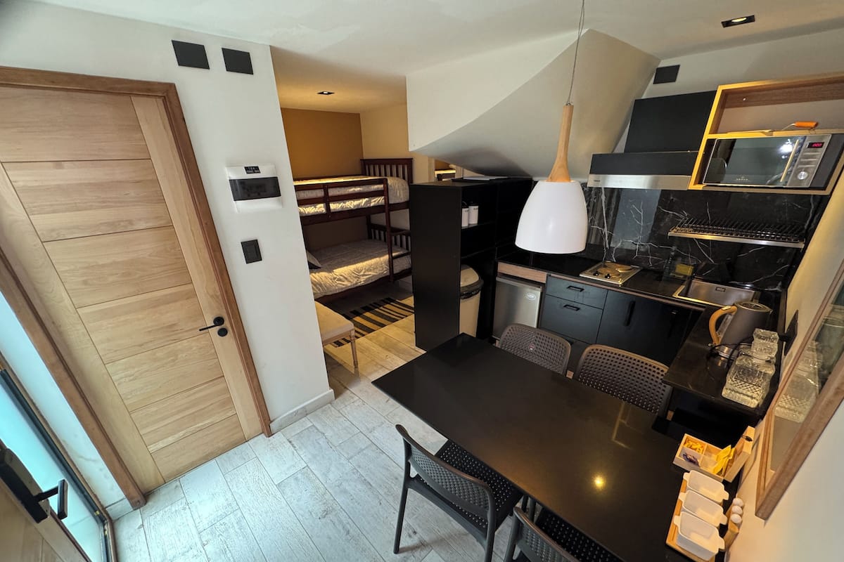 Una cocina compacta con microondas, pileta y pequeño refrigerador. Hay una mesa de comedor con sillas y un banco.