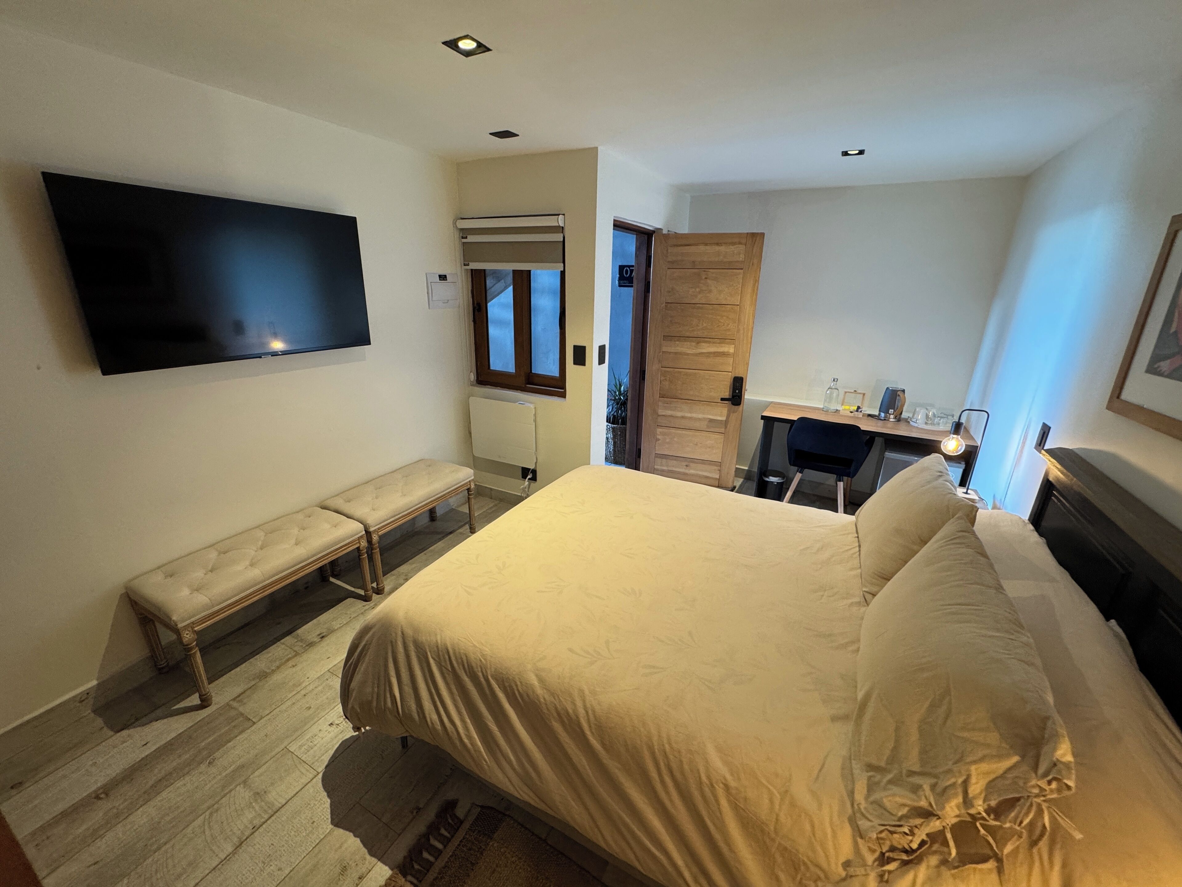 Un dormitorio con una cama, un banco, un escritorio, una silla, un televisor y una puerta.