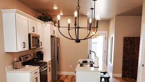 A modern kitchen with white cabinetry, stainless steel appliances, and a glass dining table surrounded by four chairs. - NEW! Sparkling 1-Level 2-BDRM King (Billings)