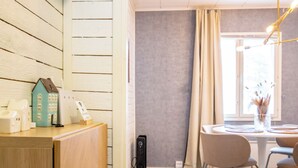 Una habitación de hotel moderna con un mostrador de recepción de madera, una pequeña decoración con forma de casa, una mesa redonda con sillas y un ventanal con cortinas.