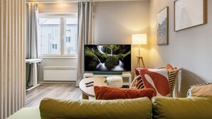Una habitación de hotel moderna con una televisión grande, un sofá cómodo, una mesita pequeña y una ventana con cortinas.