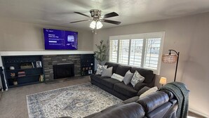 A living room with a sectional sofa, a fireplace, a TV mounted on the wall, and a ceiling fan.