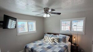 A bedroom with a bed, a ceiling fan, a TV mounted on the wall, and two windows.