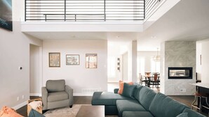 Un salon spacieux avec un plafond haut, un fauteuil gris, un canapé d’angle turquoise et un coin repas à l’arrière-plan.