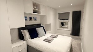 A modern bedroom with a large bed, built-in storage, and a window with curtains. - 3 bed, 'Adrian Cottage' close to Warner Bros, M25, M1. Comes with private gym (Abbots Langley)
