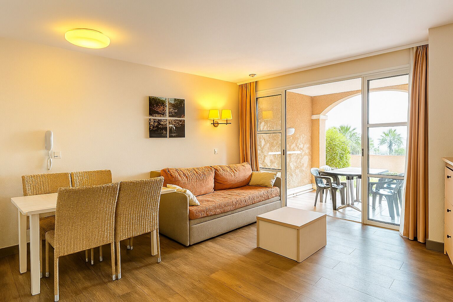 Een woonkamer met een bank, twee rieten stoelen, een kleine tafel en een glazen deur die leidt naar een buitenruimte met een tafel en stoelen.