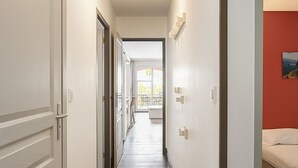 Un corridoio con pavimento in legno, pareti bianche e una parete rossa a contrasto. Ci sono porte a sinistra e un letto a destra.