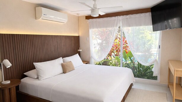Ein modernes Schlafzimmer mit einem großen Bett, einem Deckenventilator, einer Klimaanlage, einer Nachttischlampe und Blick auf Grün hinter transparenten Vorhängen.