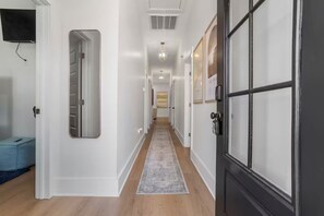 Un pasillo con paredes blancas, piso de madera y un tapete gris. Hay puertas a ambos lados, una con marco negro y la otra con marco plateado.