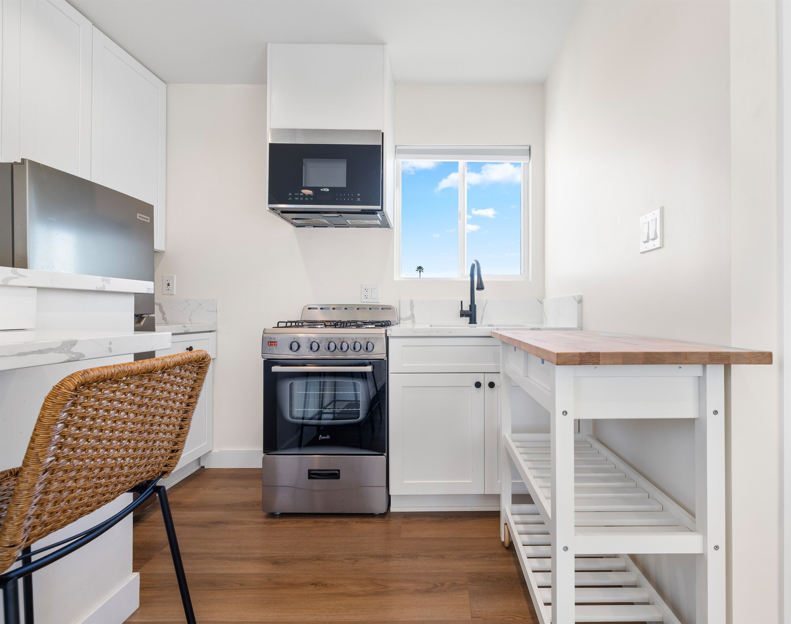 Cocina compacta con armarios blancos, horno empotrado, microondas, pileta y ventana con vista al cielo.
