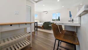 A modern room with a bed, a green sofa, a dining area with wicker chairs, a white counter, and a window with a view. - Light-Filled ADU Studio • Immaculate, Cozy & Convenient (San Diego)