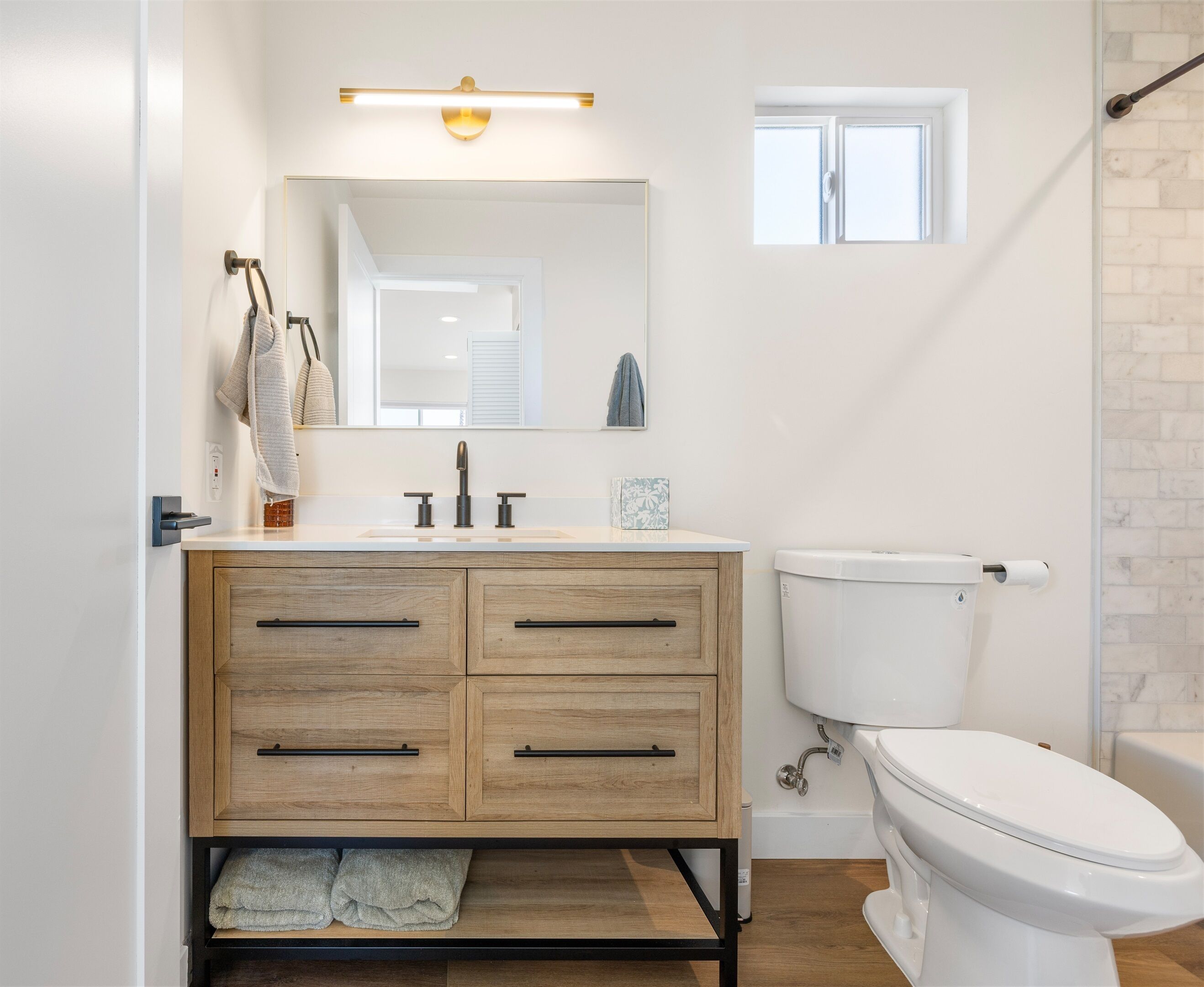 A modern bathroom with a wooden vanity, a white toilet, and a small window.