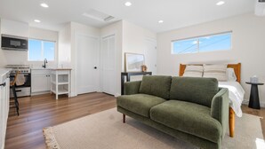 A modern, well-lit room with a kitchenette, a green sofa, a bed with white bedding, and a small side table. - Light-Filled ADU Studio • Immaculate, Cozy & Convenient (San Diego)