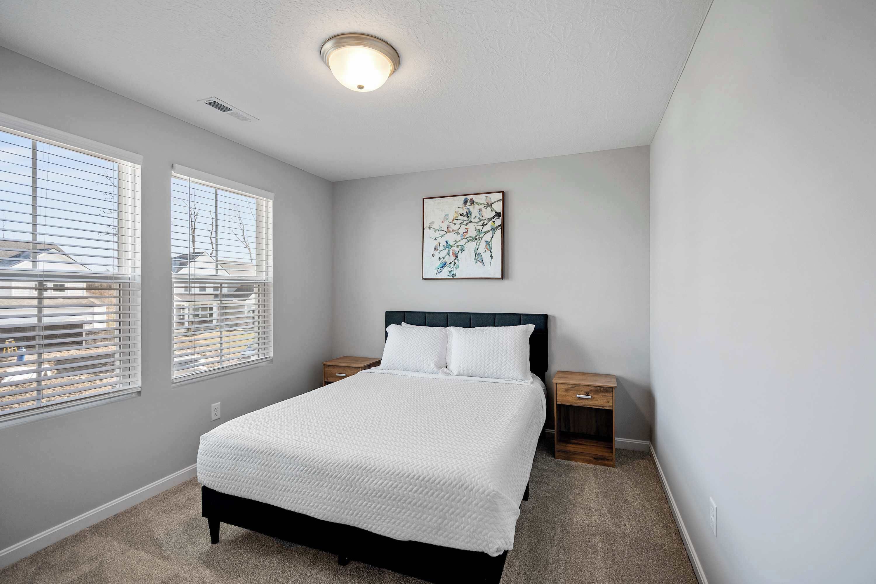 A bedroom with a bed, two bedside tables, a nightstand, a ceiling light, and a window with blinds.