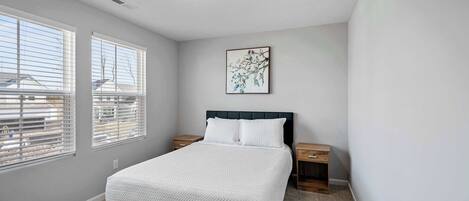 A bedroom with a bed, two bedside tables, a nightstand, a ceiling light, and a window with blinds.