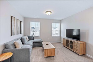 Ein Wohnzimmer mit einem grauen Ecksofa, einem Holztisch, einem Fernseher auf einem Holzständer und einem Teppichboden.