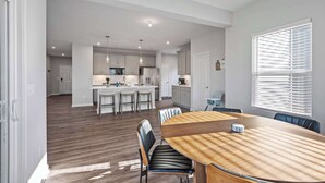 A spacious kitchen with modern appliances, a dining area with a wooden table and chairs, and a large window with blinds. - Ark & Creation Stay New Home (Walton)