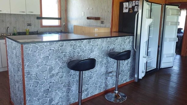 Una cocina con protector mural de piedra, dos taburetes de bar, un refrigerador y una puerta de madera.