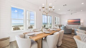 A dining area with a wooden table set for six, surrounded by cushioned chairs, a chandelier, and a view of the ocean through large windows. - NEW! Lux Beachfront, Views, Heated Pool, Sleeps 24 (Panama City Beach)