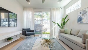 Un salon moderne avec une table basse en verre, un canapé, un fauteuil, un téléviseur fixé au mur et une porte-fenêtre coulissante donnant sur un espace extérieur.