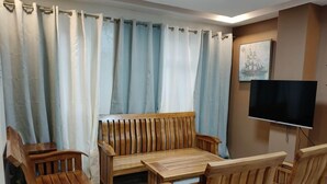Ein Zimmer mit einer Holz-Sofagarnitur, einem an der Wand montierten Fernseher und einem Fenster mit hellblauen Vorhängen.