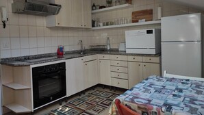 Ett kompakt kök med en inbyggd ugn, ett kylskåp och en mikrovågsugn. Det finns ett litet handfat och en bänkskiva med en vattenkokare och en vattenflaska. Köket har vita skåp och en kakelbeklädd vägg bakom bänkskivan.