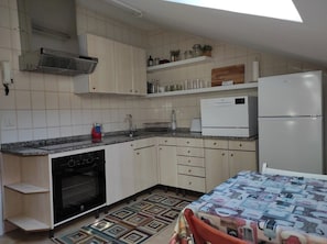A compact kitchen with a built-in oven, a refrigerator, and a microwave. There is a small sink and a countertop with a kettle and a water bottle. The kitchen has white cabinets and a tiled backsplash.