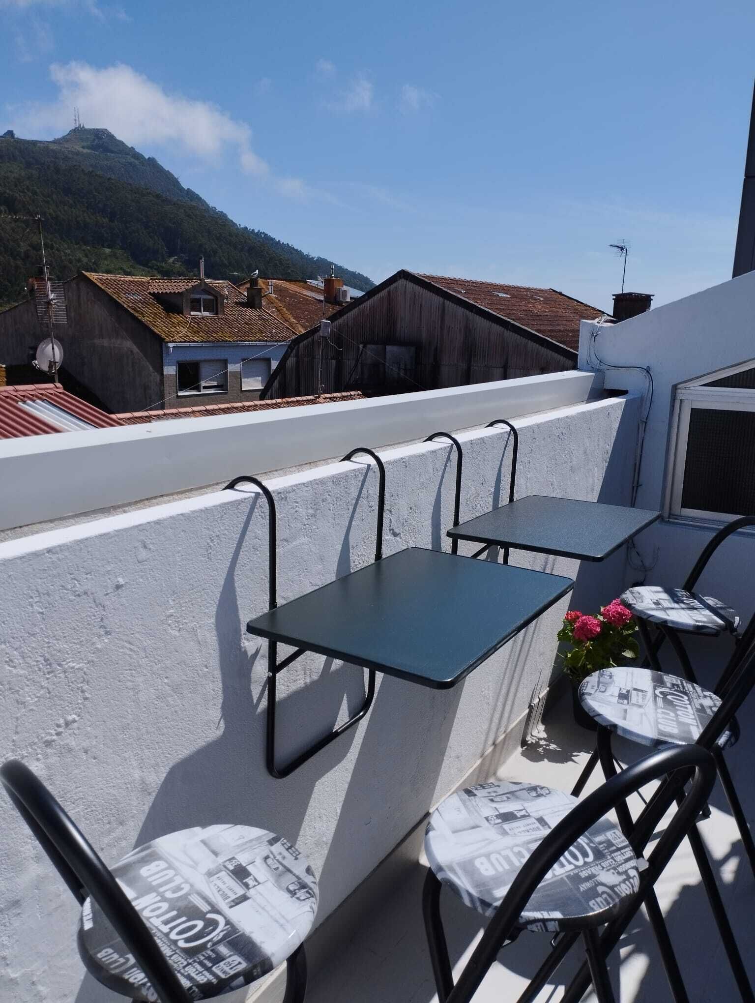 A rooftop terrace with a table and chairs, overlooking a street with buildings and a mountain in the background.