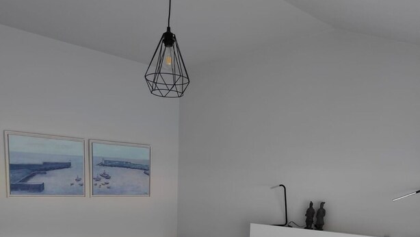 Ett sovrum med en säng, en vit sänggavel, grå sängkläder, två tavlor och en hängande lampa.