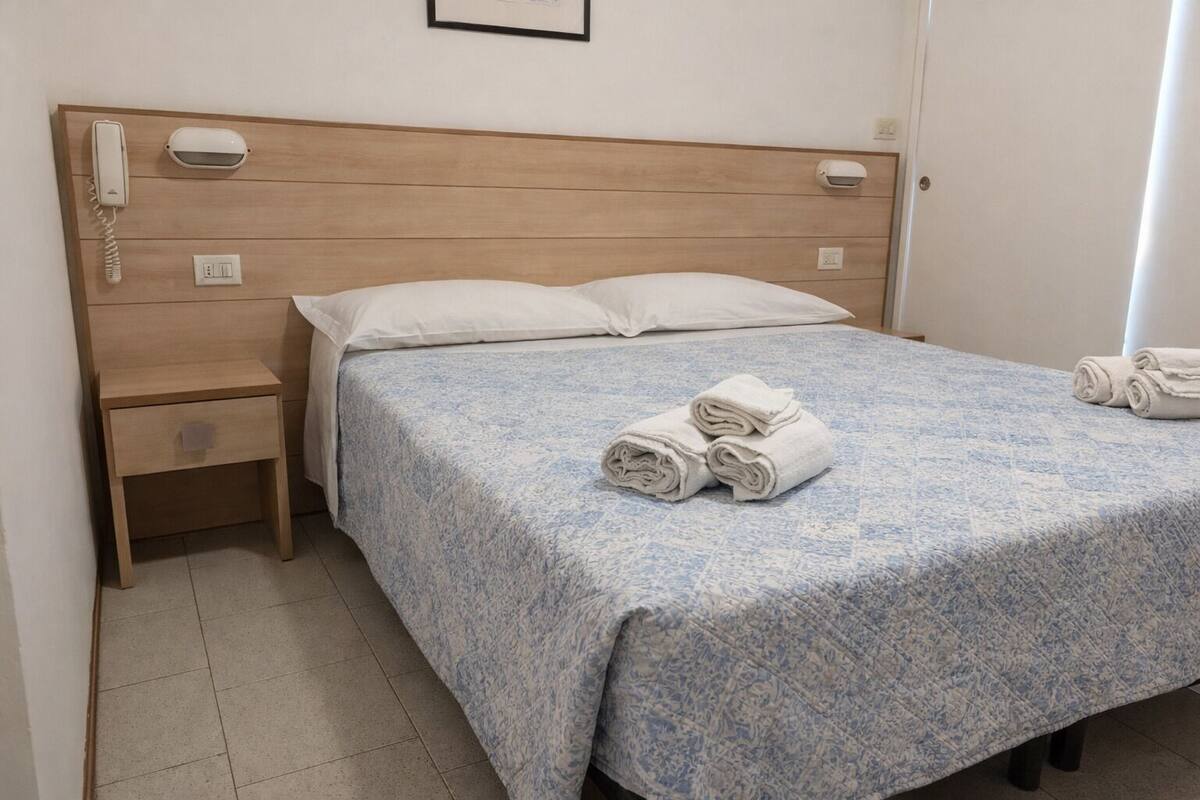 Hotellihuone, jossa on puinen päätyseinä, yksi sänky sinivalkoisella kuviollisella peitteellä, kaksi rullalle taitettua pyyhettä sängyllä, yöpöytä, jossa on puhelin ja pieni laatikko, sekä seinälle asennettu valaisin.