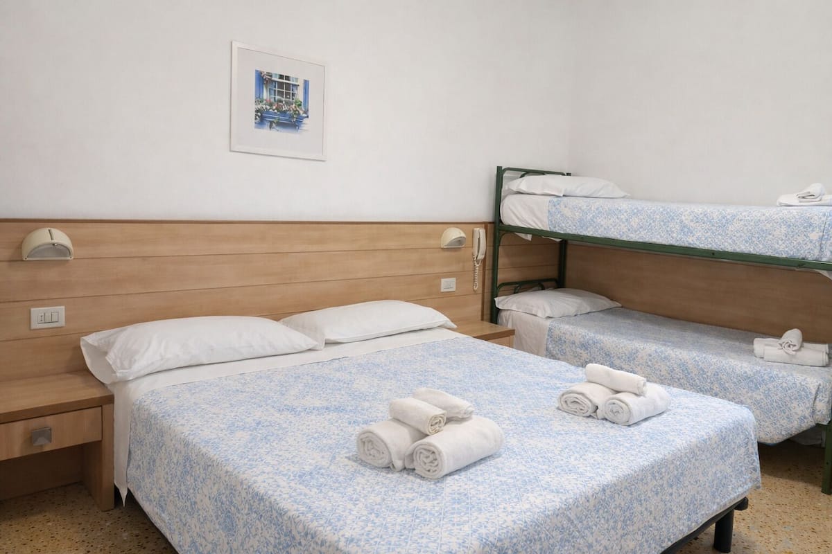 Hotellihuoneessa on kaksi sänkyä, kerrossänky, yöpöytä, lamppu, puhelin ja seinällä oleva kehystetty kuva.