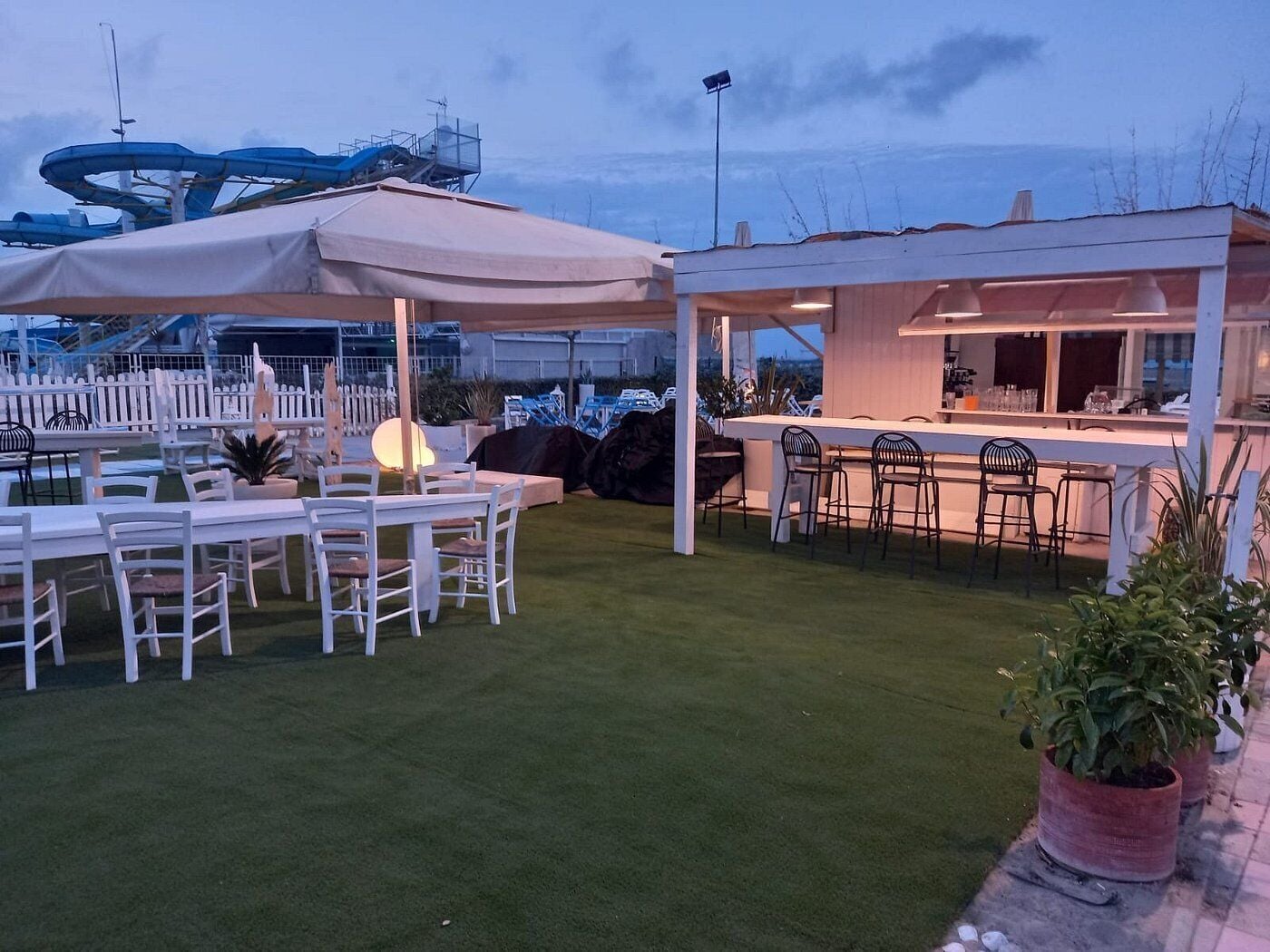 A covered outdoor dining area with tables and chairs, a bar, and a view of the sea.