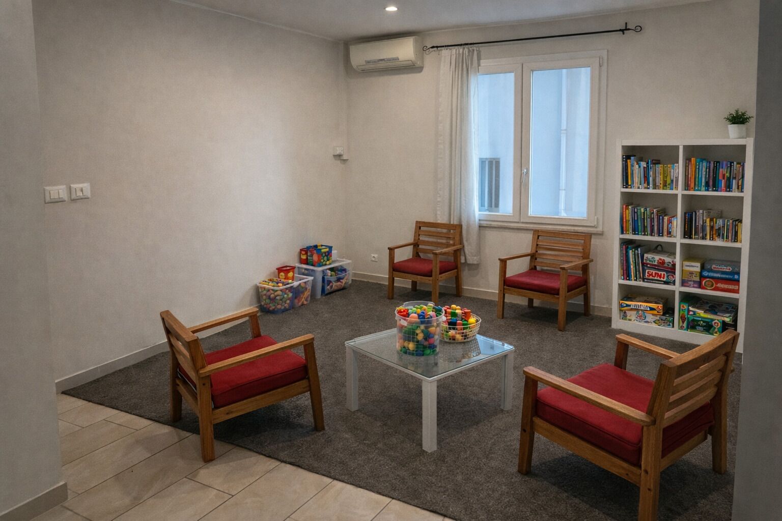 A room with a carpeted floor, a white table with toys, wooden chairs with red cushions, a bookshelf with books, and a window with curtains.
