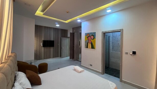 A modern hotel room with a large bed, a flat-screen TV, a painting on the wall, and a door leading to a bathroom. - LUXURY 4BED APARTMENT IKOYI (Lagos)