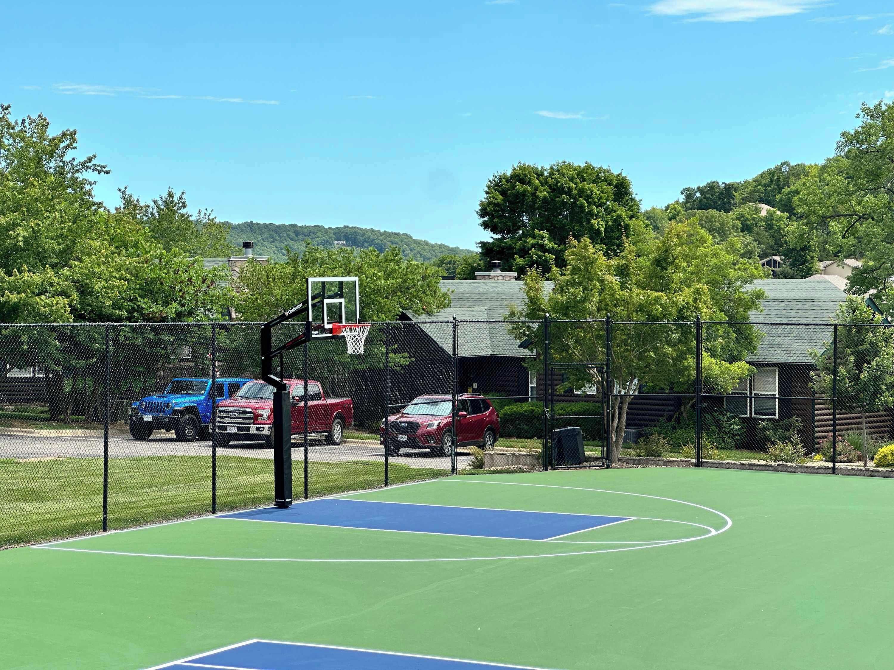 An outdoor basketball court with a chain-link fence, a basketball hoop, and parked vehicles in the background.
