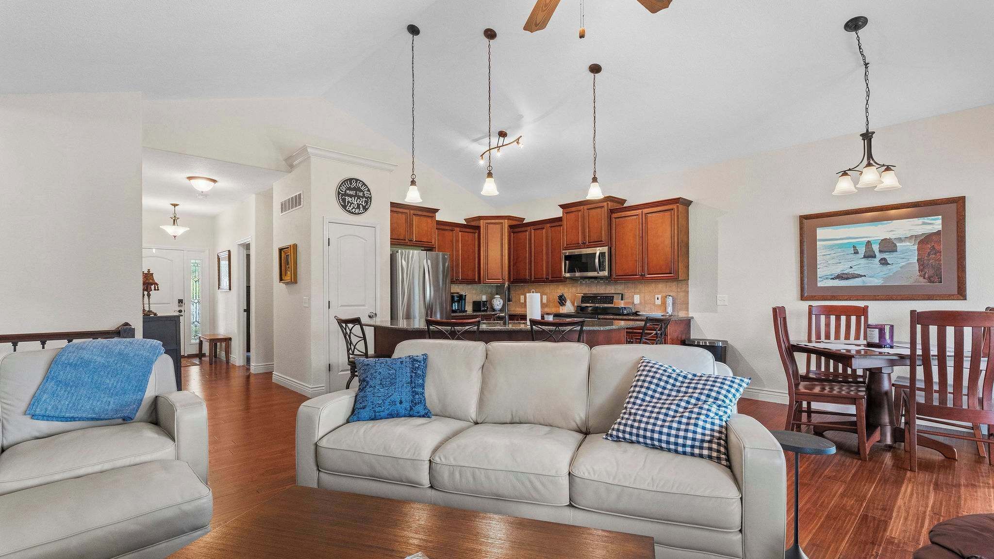 A spacious living room with a sofa, two armchairs, a dining area with a table and chairs, and a kitchen with wooden cabinets and a stainless steel refrigerator.