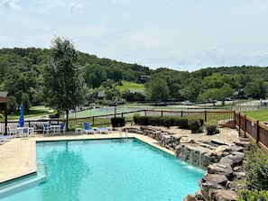 A swimming pool with a wooden deck and lounge chairs, surrounded by a wooden fence and a view of a tennis court and trees.