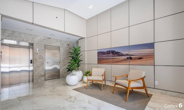 Un hall moderno con suelo de mármol, una planta en maceta grande, dos sillones, un ascensor y un cuadro de gran formato en la pared.