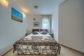A neatly made bed with a patterned bedspread, two pillows, and rolled towels on a nightstand. There is a painting on the wall, a window with sheer curtains, and a ceiling light fixture. - A 2 Comfortable apt w Terrace & Garden, Near Beach (Split)