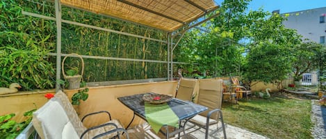 Uma área de estar coberta ao ar livre, com cadeiras e uma mesa de metal, rodeada por vegetação e um edifício ao fundo.