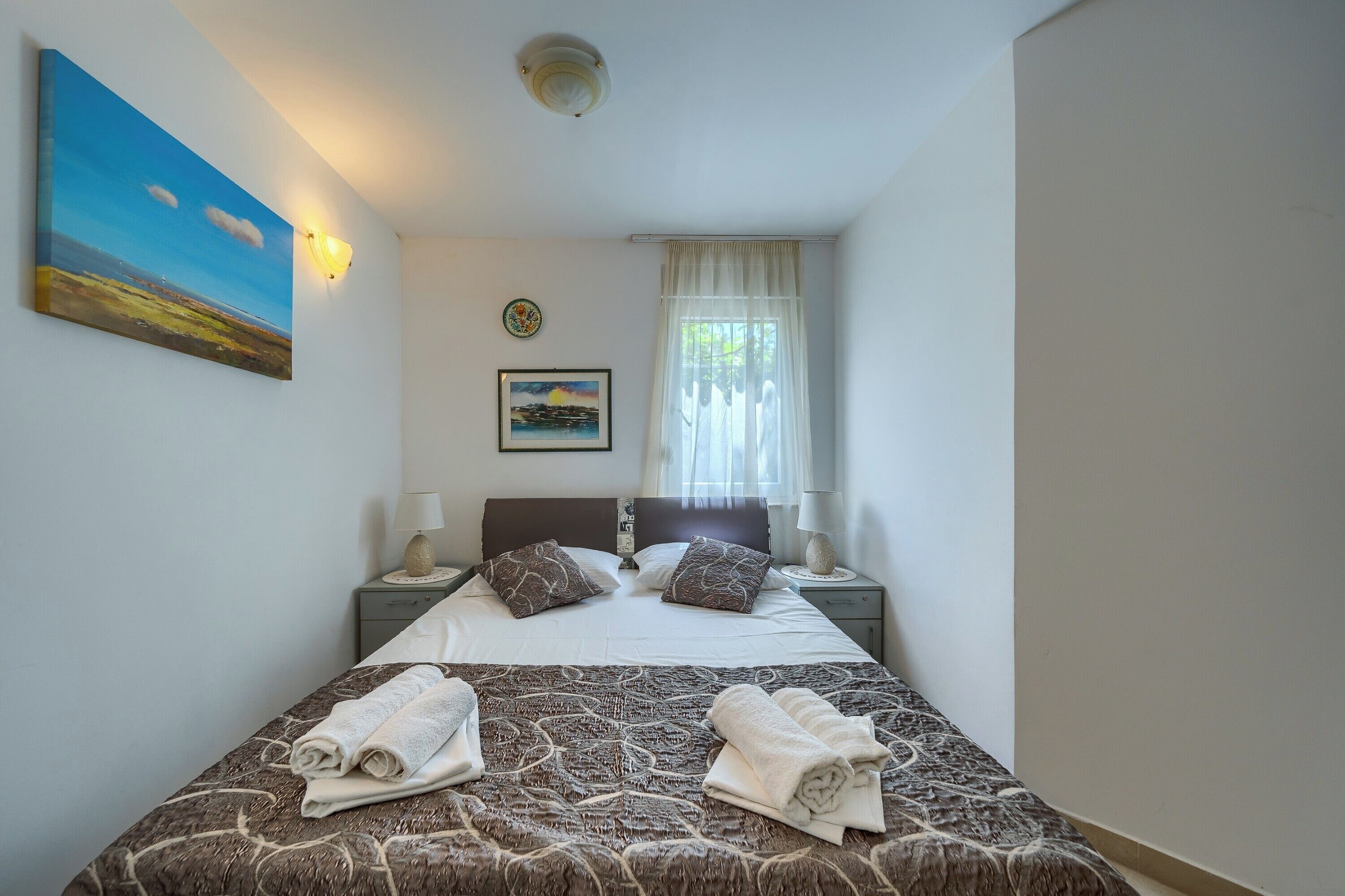 A neatly made bed with a patterned bedspread, two bedside lamps, a nightstand with a lamp, and a painting on the wall.